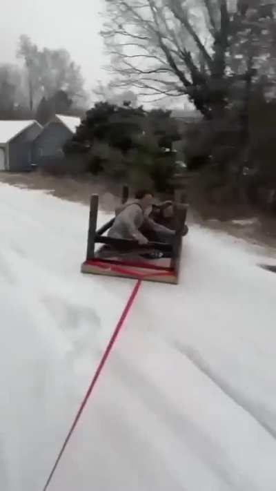 College Students using a upside down table attached to a car as a sled on a presumably icy road.